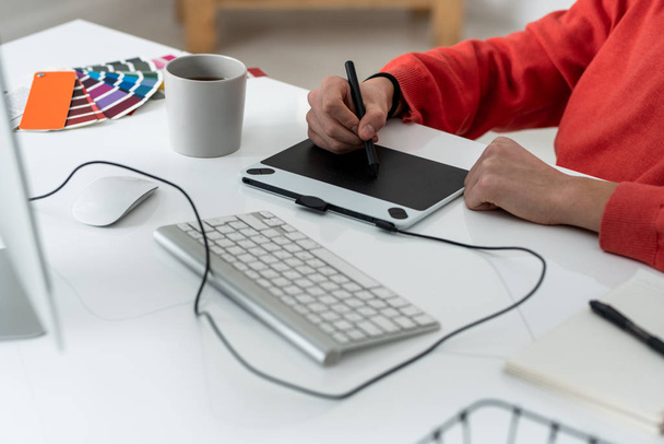 Manos de hombre joven freelancer con lápiz óptico retocando fotos en la tableta gráfica mientras está sentado junto al escritorio
 - Foto, imagen