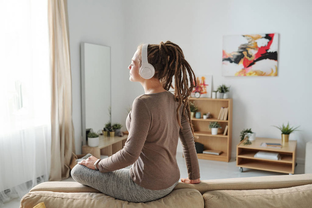 Pretty young casual female with dreadlocks enjoying meditation music in headphones while sitting on couch - Photo, image