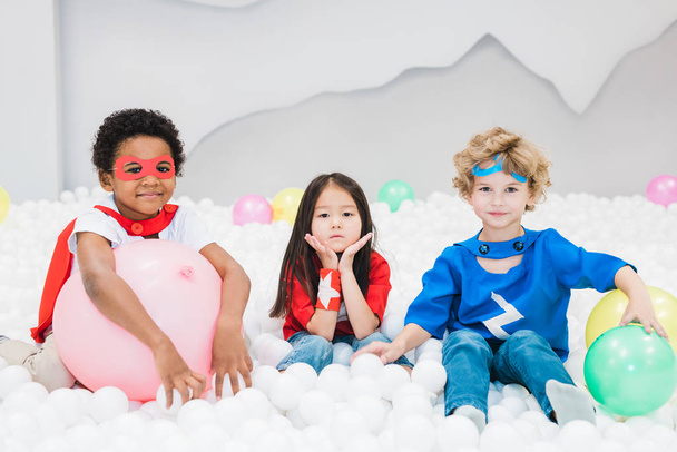 Suloisia pieniä kulttuurienvälisiä ystäviä puvuissa, jotka leikkivät valkoisilla ilmapalloilla lastenhuoneessa tai päiväkodissa
 - Valokuva, kuva