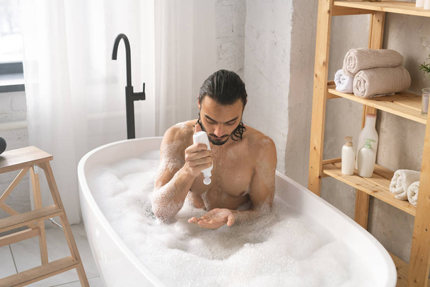 Mokrý mladý muž bez košile pomocí sprchový gel při koupeli s pěnou v koutě koupelny - Fotografie, Obrázek