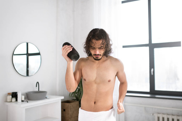 Energetic guy with white towel on hips and portable wireless bluetooth speaker dancing in bathroom - Foto, Imagen