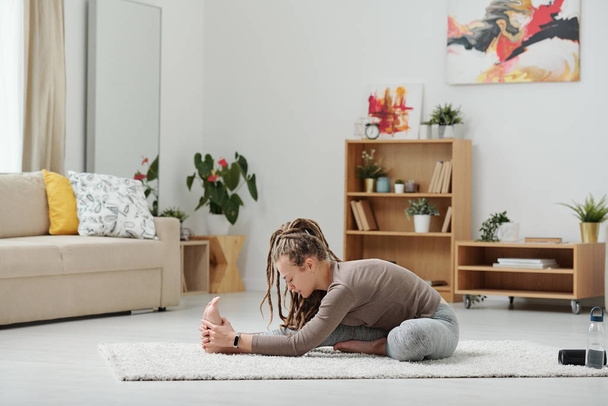 Активна дівчина сидить на підлозі і розтягує ногу вперед під час фізичних вправ в домашньому середовищі на дозвіллі
 - Фото, зображення