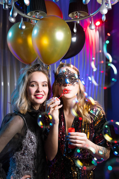 Menina bonita em renda máscara veneziana soprando bolhas de sabão com seu amigo de pé perto em festa no clube noturno - Foto, Imagem