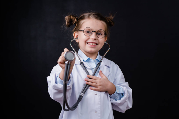 Милая веселая маленькая девочка в белом халате, держащая стетоскоп перед камерой, стоя на черном фоне
 - Фото, изображение