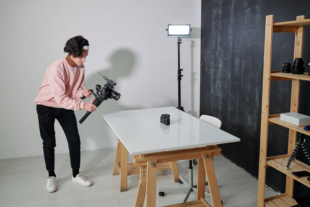 Joven video blogger en ropa de casualwear disparando nuevo equipo fotográfico mientras está de pie junto al escritorio en el estudio
 - Foto, imagen