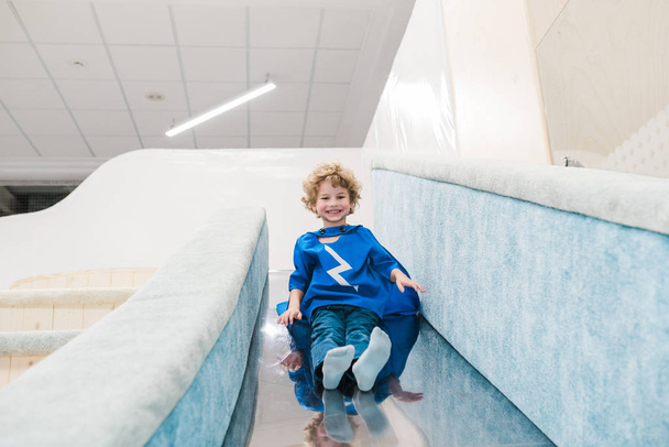 Mignon petit garçon joyeux en costume bleu de Superman descendant glissière de chute sur l'aire de jeux dans le centre moderne des enfants
 - Photo, image