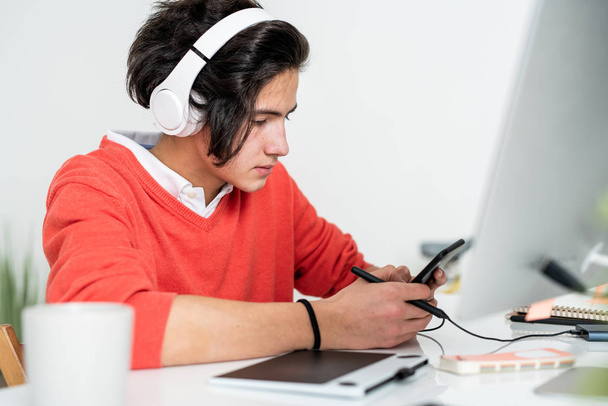 Cara ocupada com fones de ouvido rolando no smartphone enquanto procura informações na frente do computador
 - Foto, Imagem