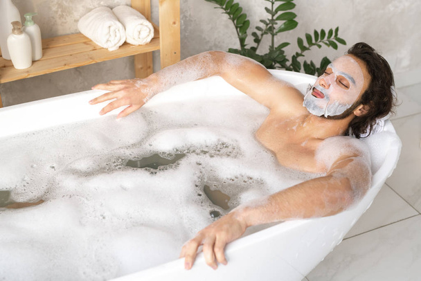 Serene young man with face mask relaxing in hot bath with foam while keeping his eyes closed - 写真・画像