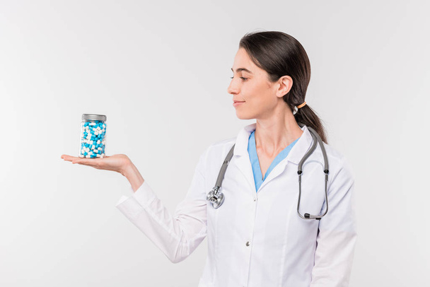 Jolie jeune femme clinicienne ou infirmière dans un bocal de whitecoat avec des pilules à portée de main tout en se tenant dans l'isolement
 - Photo, image