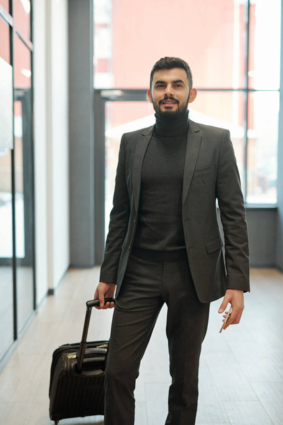 Joven hombre de negocios con maleta y smartphone de pie delante de la cámara mientras espera el anuncio de salida en el aeropuerto
 - Foto, imagen