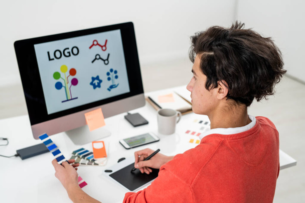 Hombre joven con paleta de colores creando el logotipo para el sitio web mientras está sentado frente a la computadora y el uso de gráficos tableta
 - Foto, imagen