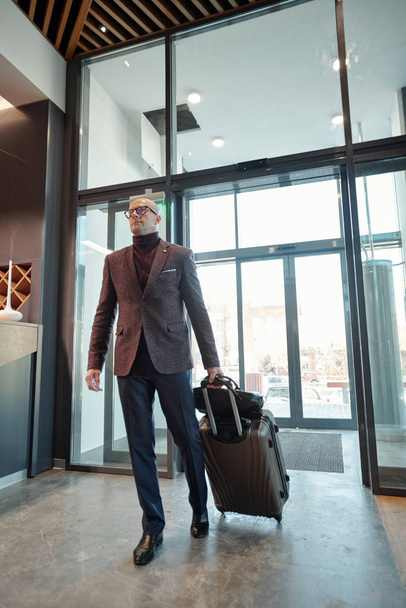 Ältere männliche Geschäftsreisende ziehen Koffer beim Betreten der Hotellounge nach der Ankunft im Ausland - Foto, Bild