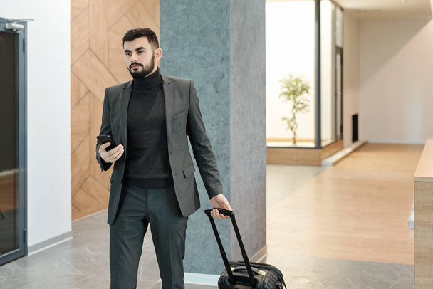 Молодой элегантный бизнесмен вытягивает чемодан, двигаясь по гостиной отеля и ожидая администратора
 - Фото, изображение