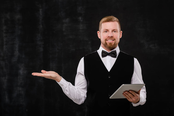 Happy young bearded waiter with digital tablet showing you where to go while standing in front of camera - Photo, Image