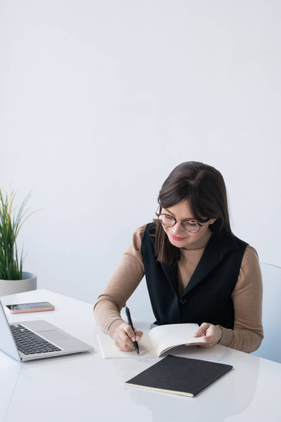 Молода успішна бізнес-леді записує план робочого дня, сидячи за столом перед ноутбуком
 - Фото, зображення