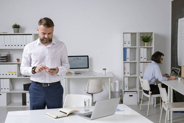 Успешный бизнесмен в формальной одежде прокручивая в смартфоне для контакта, стоя за столом в офисной среде
 - Фото, изображение