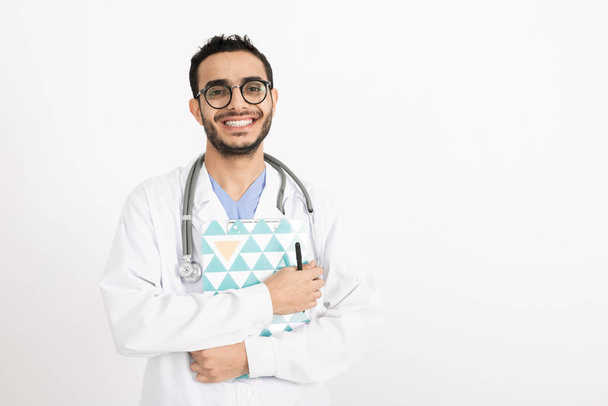 Stetoskobu, not panosu ve kalemi olan mutlu, başarılı genç klinisyen yalnız başına kameranın önünde duruyor. - Fotoğraf, Görsel