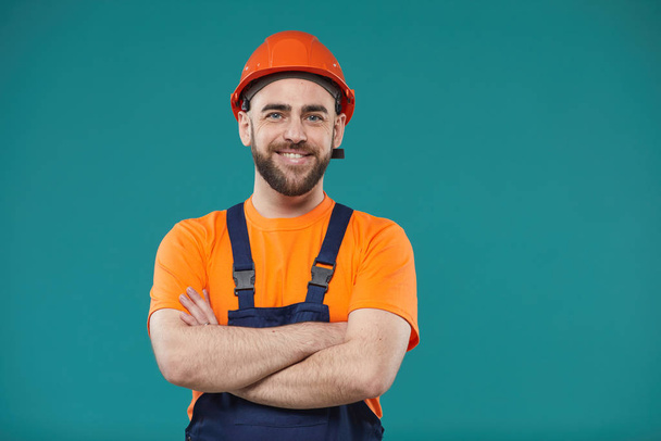 Horizontal waist up studio portrait of cheerful man wearing hardhat and workwear standing with atms crossed looking at camera - 写真・画像