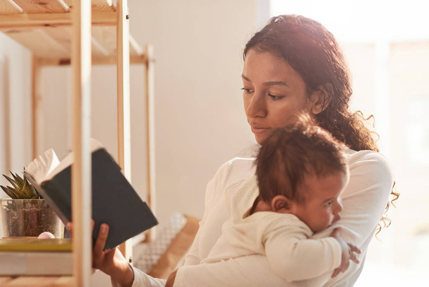 Warm-toned portrait of young African-American mother reading bonk and cuddling cute baby, copy space - Photo, Image