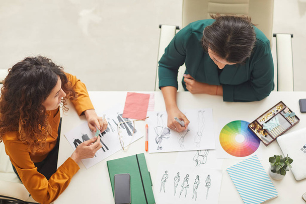 Twee stijlvolle jonge vrouwen creëren mode ontwerp schetsen zitten samen aan kantoor tafel, horizontale top vew shot - Foto, afbeelding