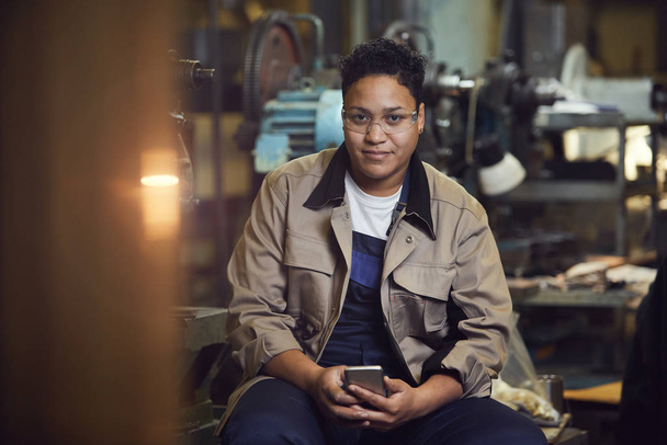 Портрет современной женщины смешанной расы, улыбающейся в камеру, позируя в заводской мастерской
 - Фото, изображение