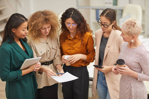 Ομάδα πέντε γυναικών που μιλούν για τη δουλειά κατά τη διάρκεια του διαλείμματος καφέ στέκονται μαζί στο γραφείο - Φωτογραφία, εικόνα