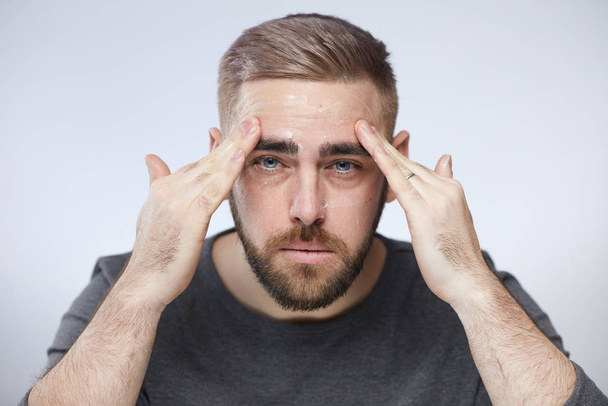 Ritratto orizzontale di testa e spalle di giovane uomo che applica crema nutriente per il viso sul suo viso guardando la fotocamera, sfondo bianco
 - Foto, immagini