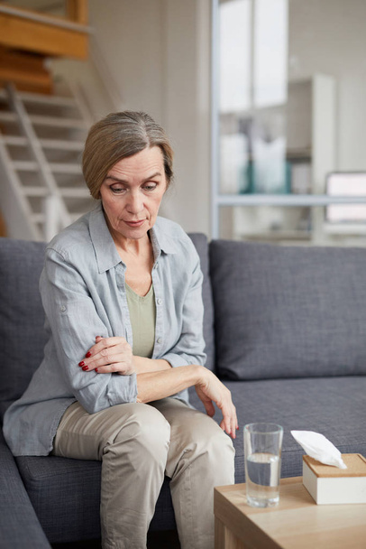 Portrait of sad mature woman sitting on couch at home and looking down with sad face expression, tissues and water on table, copy space - Photo, Image