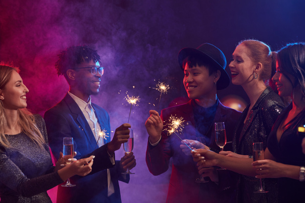 Grupo multi-étnico de jovens elegantes que iluminam faíscas durante a festa de Natal no clube noturno fumegante
 - Foto, Imagem