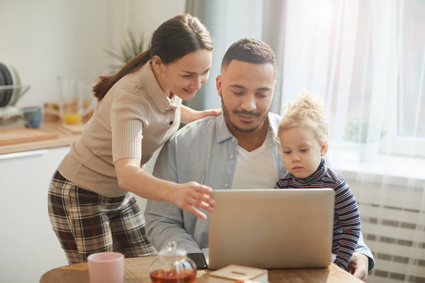 Modern melez ailenin sıcak tonlu portresi bilgisayarı kullanırken küçük tatlı kızıyla rahat bir mutfakta oturuyor. - Fotoğraf, Görsel