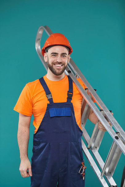 Studio portrait of young adult male electrician holding portable ladder looking at camera smiling - Foto, imagen
