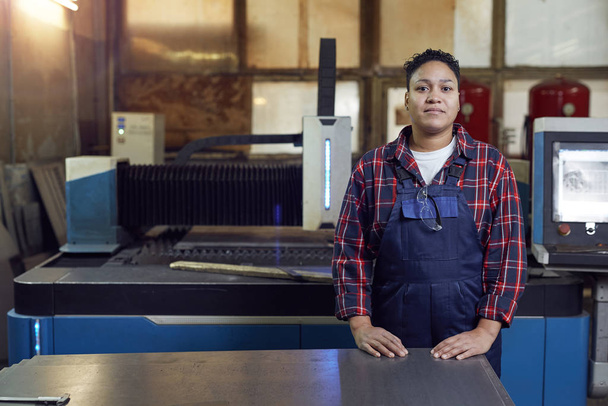 Derékig érő portré vegyes fajú női munkásról, ahogy a műhelyben álló ipari gépegységeknél álló kamerát nézi, másolja a helyet - Fotó, kép
