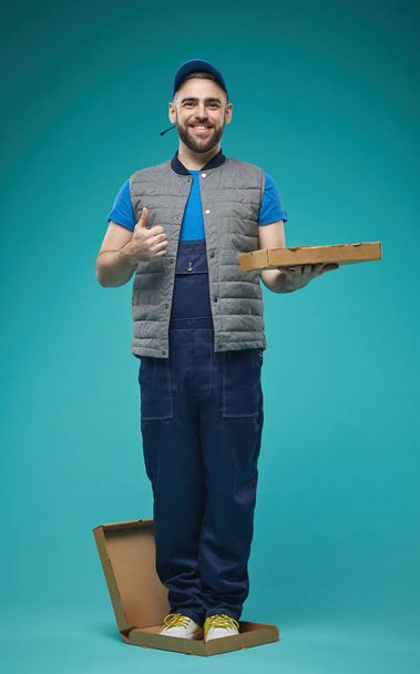Κάθετη full shot στούντιο πορτρέτο του αστείου διανομέα στέκεται σε κουτί πίτσα δείχνει τον αντίχειρα μέχρι χαμογελώντας στην κάμερα - Φωτογραφία, εικόνα