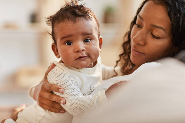 warmgetöntes Porträt des süßen afrikanisch-amerikanischen Babys, das in die Kamera schaut, während es mit der jungen Mutter zu Hause kuschelt, Kopierraum - Foto, Bild