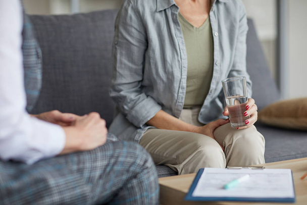 Cropped portrait of adult woman holding glass of water while sharing troubles with psychologist during therapy session, copy space - Photo, image