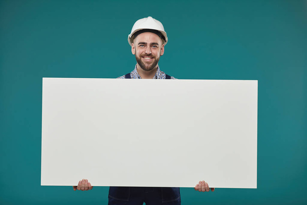 Horizontal studio portrait shot of happy young adult manual worker holding blank poster looking at camera smiling, blue turquoise background - 写真・画像