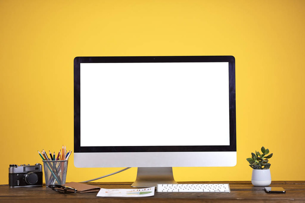 薄い黄色の背景の上に木製の机で空白の白いコンピュータ画面と職場の概念、コピースペース - 写真・画像