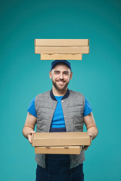 Vertical studio portrait of food delivery guy with three pizza boxes on head holding another three boxes in hands looking at camera smiling - Photo, Image