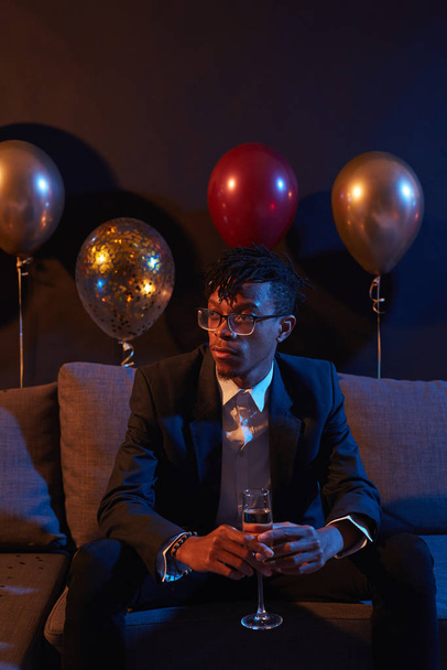 Ritratto di uomo afro-americano alla moda che tiene un bicchiere di champagne seduto sul divano in una stanza scarsamente illuminata al night club, copia lo spazio
 - Foto, immagini