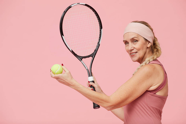 Pas w górę portret aktywnej dojrzałej kobiety trzymającej rakietkę tenisową stojąc na pastelowym różowym tle, przestrzeń do kopiowania - Zdjęcie, obraz