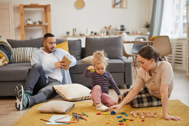 Retrato de larga duración de la familia moderna de raza mixta jugando con una linda niña en el acogedor interior de la sala de estar, espacio de copia
 - Foto, Imagen