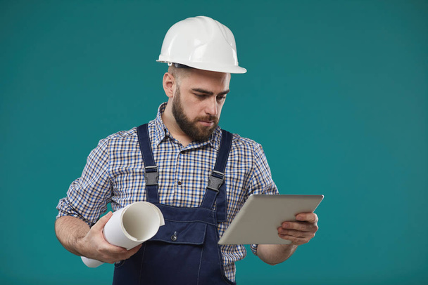 Medium studio portrait of Caucasian engineer standing against blue turquoise wall holding building plan and using tablet computer - Photo, Image
