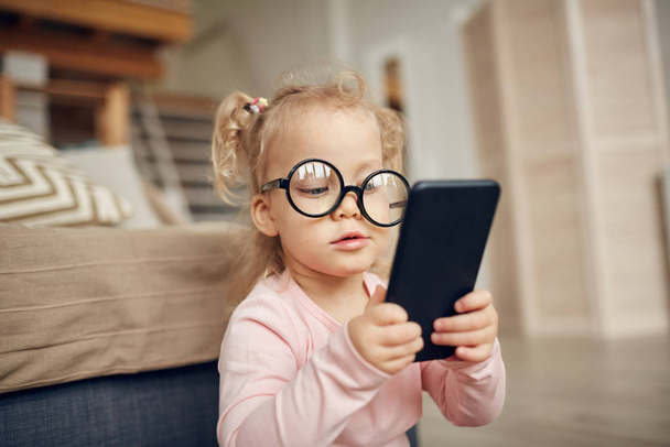 Retrato de niña linda con gafas grandes y plying con teléfono inteligente mientras está sentado en el suelo en el acogedor interior del hogar, espacio de copia
 - Foto, imagen