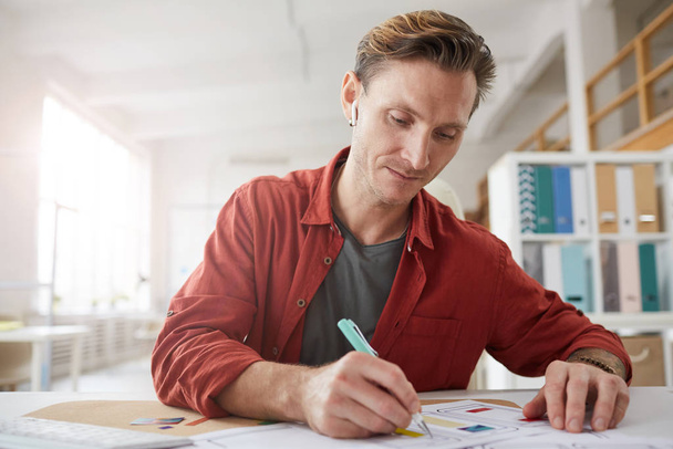 Portret współczesnego dorosłego mężczyzny robiącego notatki podczas pracy w biurze i słuchającego muzyki przez bezprzewodowe słuchawki, przestrzeń do kopiowania - Zdjęcie, obraz