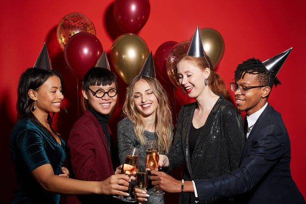 Πολυεθνική ομάδα χαρούμενων νέων που κλίνουν ποτήρια σαμπάνιας απολαμβάνοντας το κόμμα στέκεται πάνω από το κόκκινο φόντο με μπαλόνια, αντιγραφή χώρου - Φωτογραφία, εικόνα