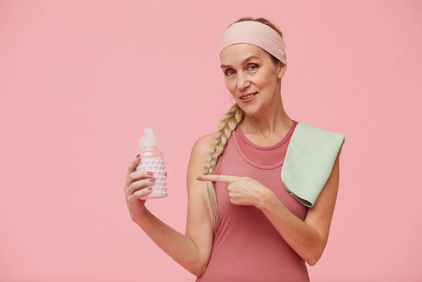 Portrait taille haute de femme mature en forme regardant la caméra tout en pointant vers la bouteille d'eau sur fond rose pastel, espace de copie
 - Photo, image