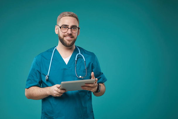 Horizontal medium studio portrait of Caucasian medical worker wearing blue uniform holding tablet computer looking at camera smiling - Fotoğraf, Görsel