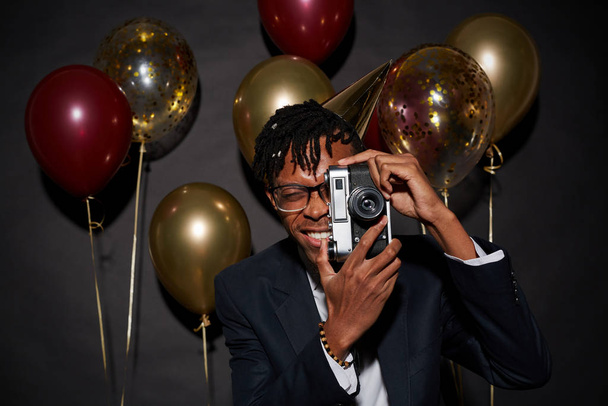 Cintura até retrato de homem africano contemporâneo segurando câmera fotográfica enquanto posando contra fundo preto com balões de festa, tiro com flash
 - Foto, Imagem