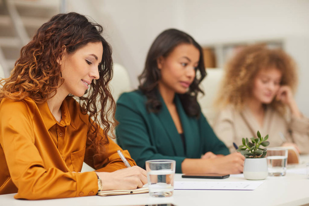 Horizontale oogfoto van drie stijlvolle jonge vrouwen die notities maken tijdens zakelijke bijeenkomst in modern kantoor - Foto, afbeelding