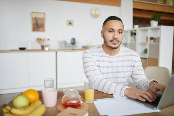 Portrait of handsome mixed-race man looking at camera while working from home and using laptop at kitchen table, copy space - Fotografie, Obrázek